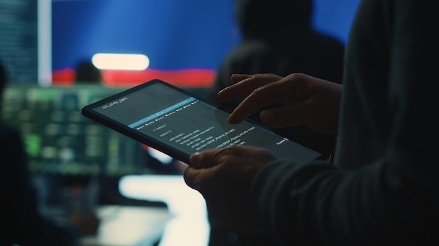 A cybersecurity professional analyzing network data on a tablet in a dark security operations center, with multiple screens displaying real-time system monitoring.